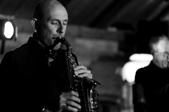 A musician plays a saxophone with their eyes closed with another musician in the background.