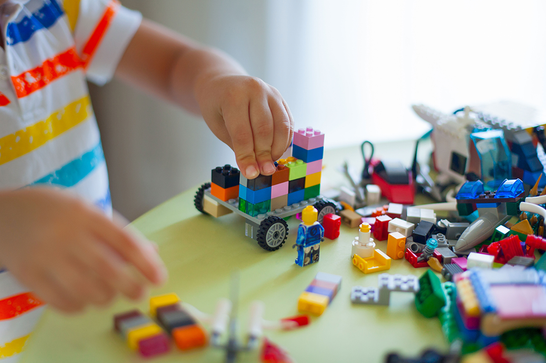 The hands of a child, who is wearing a brightly coloured striped t shirt, building a vehicle from LEGO bricks with more  LEGO pieces and a LEGO person scattered around.