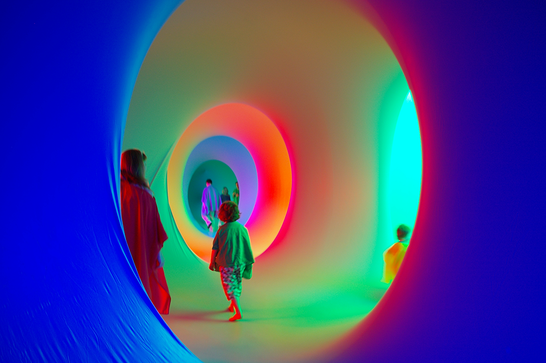 Framed through a blue circular entrance, an adult and two children explore a colour circular chamber and you can see more similar chambers of different colours in the background with another family in the distance.
