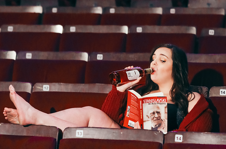 A woman with black hair in a red top sits in a theatre with her feet up, drinking from a bottle of wine and reading a book.
