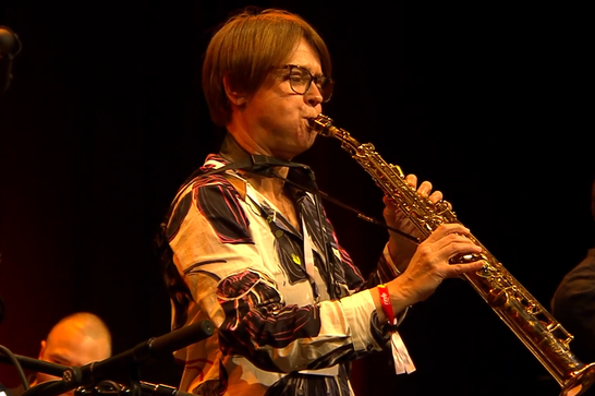 A musician with brown hair and glasses playing a soprano saxophone.