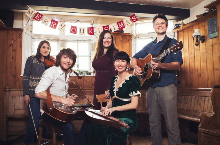 Group of five musicians with their instruments. The two people at the front are sat, and the other three are stood behind them. Hanging behind them is bunting with letters that spell out 'Merry Christmas'.