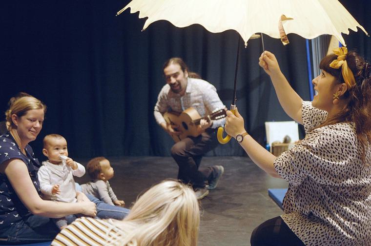 Woman on the left with baby on her lap. Man in the middle is knelt down playing the guitar. A baby is starting to crawl towards him. Woman on the right hold up a big yellow umbrella and sings. She has a yellow bow in her hair.