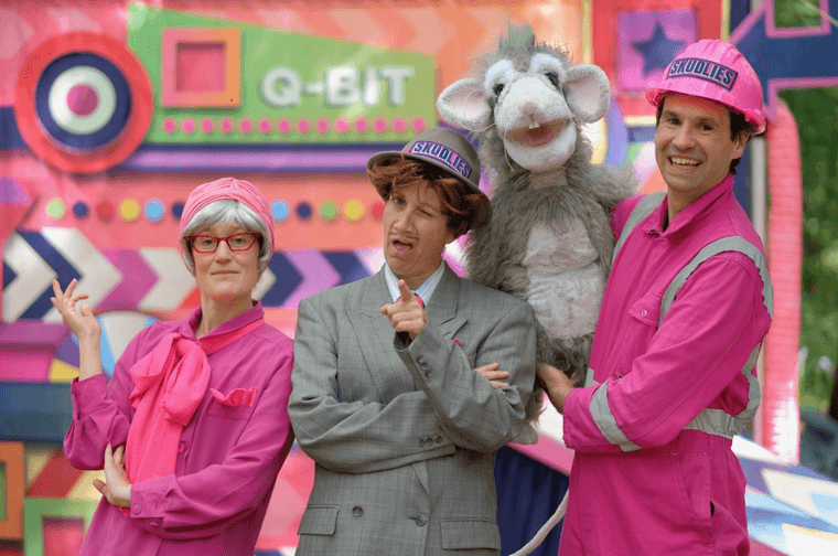 Three people stood in a line. The person in the middle is wearing a grey suit and is winking. The other two people are wearing pink hazard suits and hard hats. The man in the left is holding a large rat puppet.