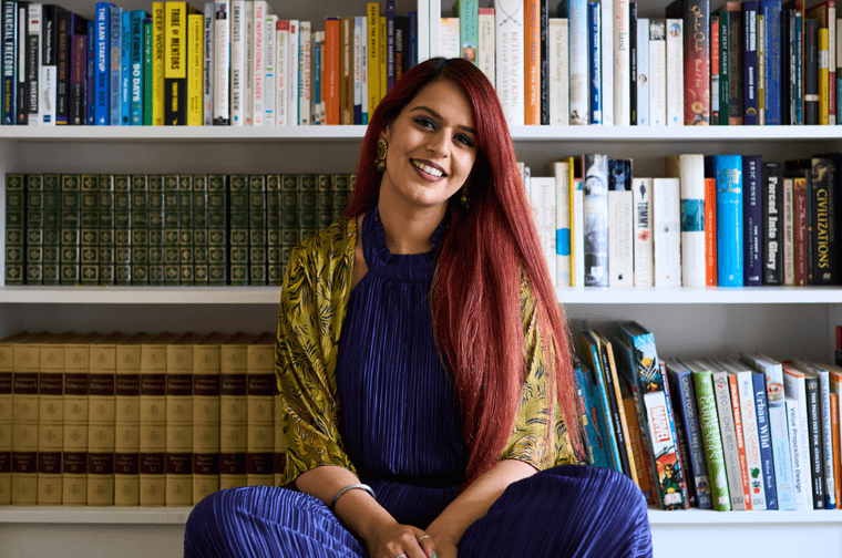 Photograph of Jaspreet Kaur. She is sat on the floor with her legs crossed, in front of a large bookcase.