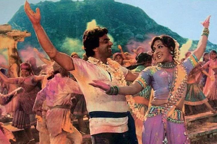 A still from the 1975 Indian film Sholay. In the picture there is a man and a woman dancing with their arms int he air. There are wearing colourful clothes. They are at Holi Festival so they are covered in coloured powder. There are lots of people behind them who are also dancing and throwing the powder.