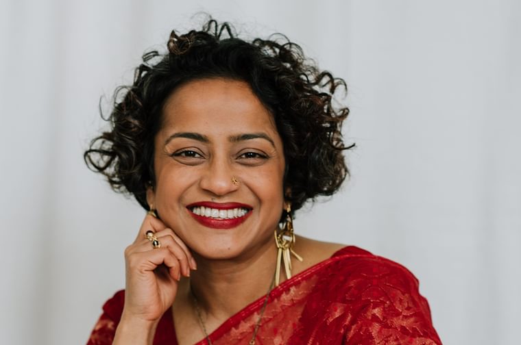 A picture of Poppy Jaman, against a white background. Poppy has short curly brown hair and is wearing a red top.