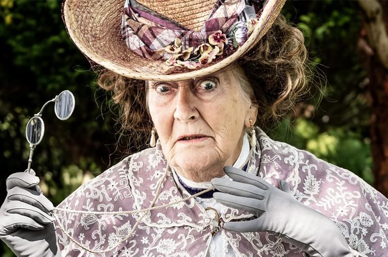 Close up photograph of an older woman looking very startled. She is wearing the clothes of an 1980s woman, with a big straw hat, gloves and violet cape. She is holding her glasses in her right hand.