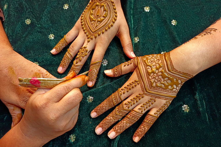 Hands decorated with intricate henna patterns on green fabric with golden embroidery, showing traditional mehndi art being applied with a cone applicator.