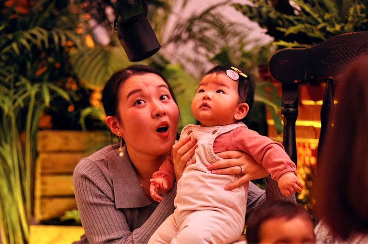 Woman holding up her baby. They are both looking up . They look excited and amazed. Behind them is a tent filled with plants and lights, to make it look like a magical rainforest.