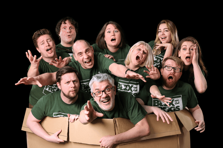 Group of ten people stood in a large cardboard box. They are all wearing green tops wit 'Box of Frogs' written on them. They are making exaggerated facial expressions and acting.