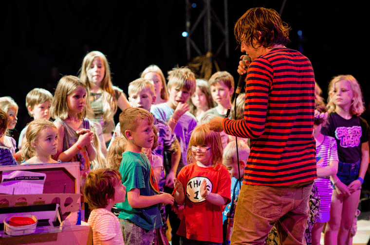 Man in red and black stripped t shirt singing on a stage. Group of children around him dancing along.