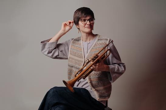 Laura Jurd is sitting on a stool, holding a trumpet. Laura has short brown hair and is wearing glasses and a beige vest top.