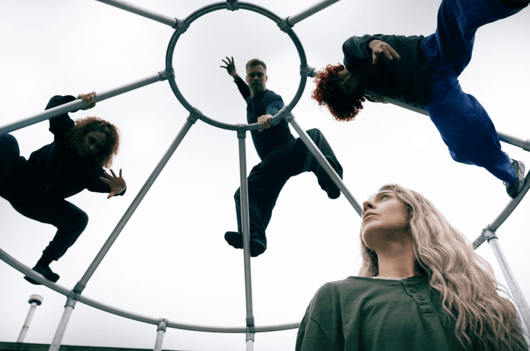 Three people climbing on a metal “birdcage" and dancing. Blow the cage is a blonde woman looking up at the other performers.