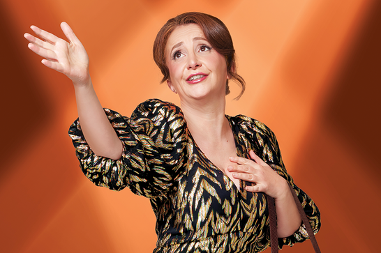 A woman wearing a gold and black dress (Lucy Porter) stands in front of an orange lit background with her right hand outstretched and her left hand to her chest.