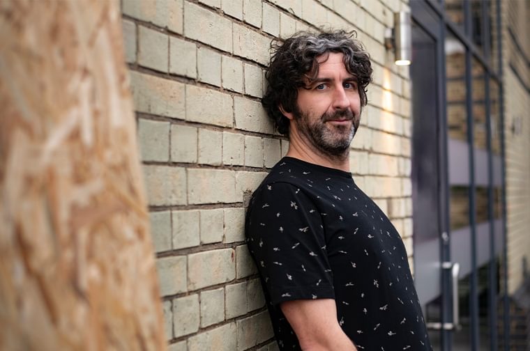 Man leaning against a brick wall. He had dark curly hair and a short beard. He is wearing a short sleeve black t shirt with a small white pattern.