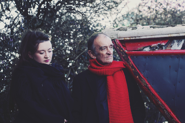 Young woman and older man in dark coats stand beside a weathered red boat against a backdrop of white flowering branches.