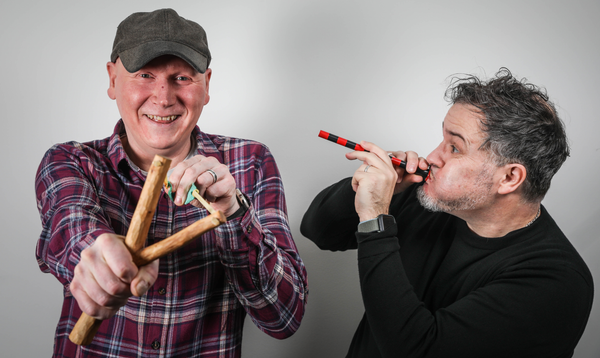 Two men against a grey background. The man on the left wears a grey cap and red plaid shirt, smiling broadly while holding a wooden sling shot. The man on the right has curly grey hair and wears a black shirt, playing what appears to be a wooden flute or pipe with red and black striping.