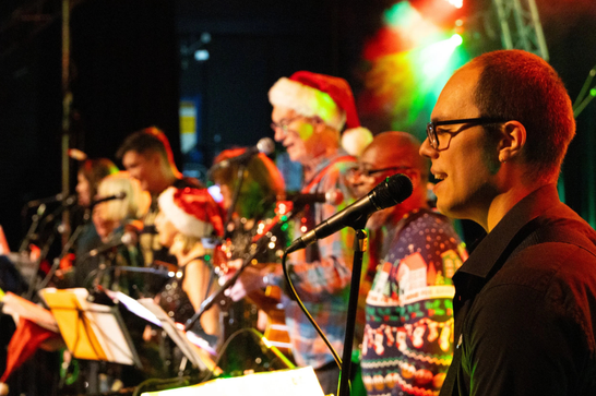A group with a variety of instruments, wearing red and white Christmas hats on a warmly lit stage.