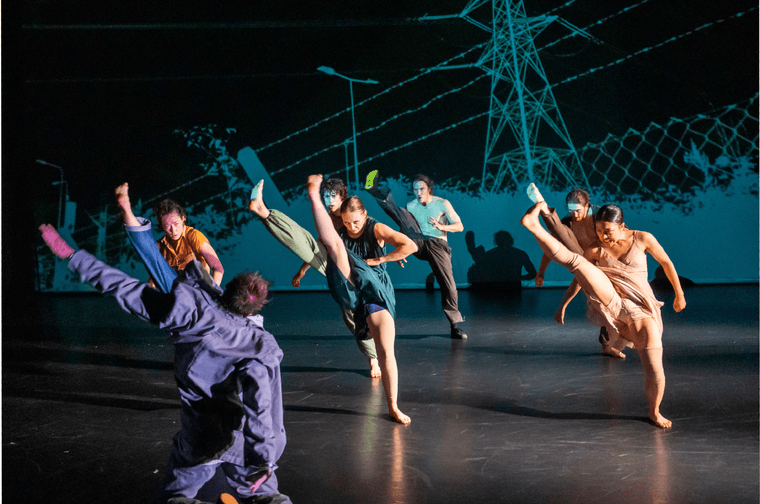 Group of people dancing on a stage. The are all kicking there right leg up in sync. Behind them is a large blue cut out in the shape of power lines and a wire fence.