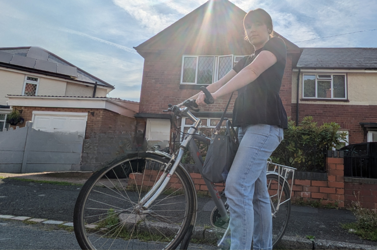 Woman wearing black ruffle top and jean holding her bike. She is stood in front of a semi-detached house and the sun is very bright behind her.