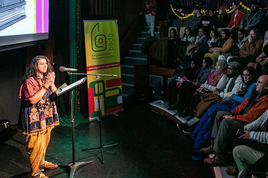 A woman in traditional South Asian attire speaks at a podium to a seated audience in a theatre setting.