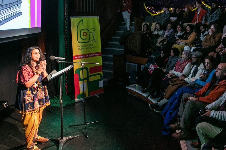 A woman in traditional South Asian attire speaks at a podium to a seated audience in a theatre setting.