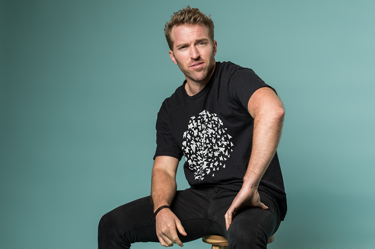 A man in a black t shirt sits on a stool against a teal background, one arm rests on his knee and he looks ahead.