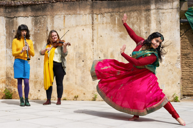 A woman in a pink and green sari performs a traditional dance on stage while two musicians play instruments in the background.
