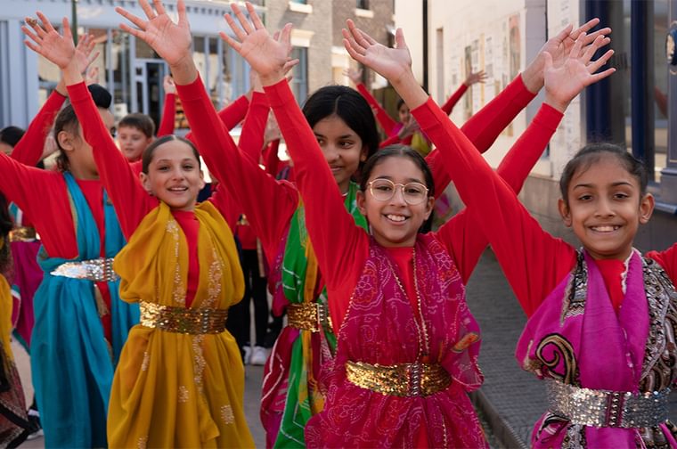 A group of young girls with their arms in the air. They are all wearing red long sleeve tops, with a sari around their necks and secured with a belt. They are smiling.
