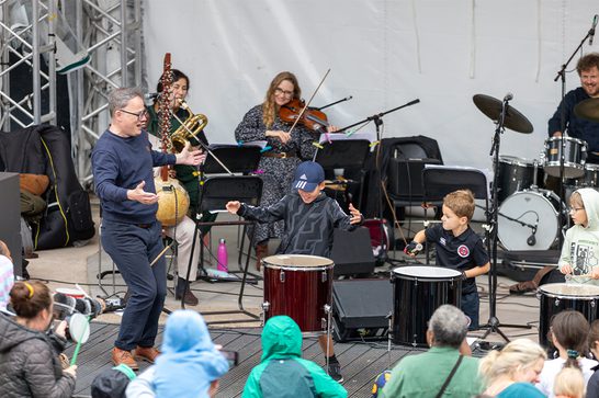 Three young boys playing large drums on stage. They are guided by a man with glasses. Behind them is a small orchestra. The backs of the heads of the audience are at the bottom of the image.