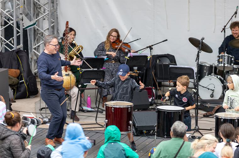Three young boys playing large drums on stage. They are guided by a man with glasses. Behind them is a small orchestra. The backs of the heads of the audience are at the bottom of the image.