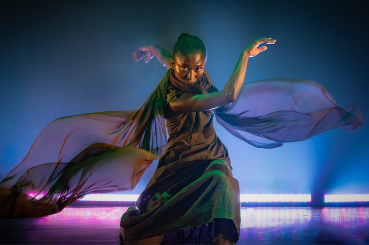 Woman in black floaty dress dancing on a stage. The stage is dark, with purple and blue lights across the bottom.