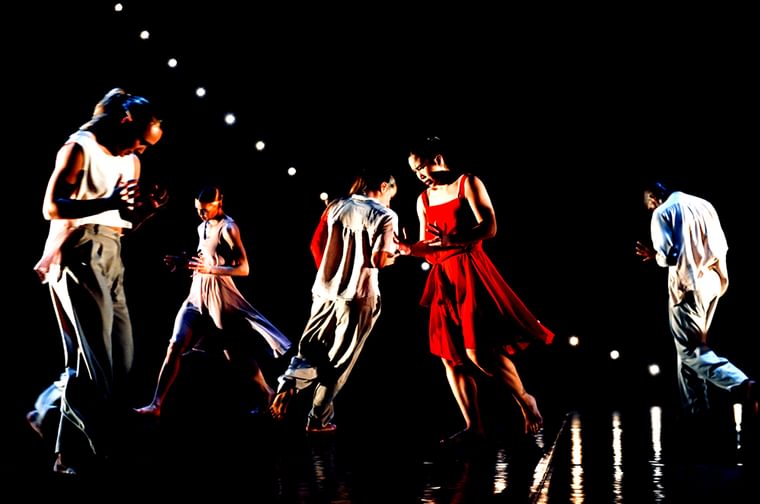Group of five people dancing on a dark stage. Four of them are wearing white loose fitting cloths and the one woman is wearing a bright red dress.
