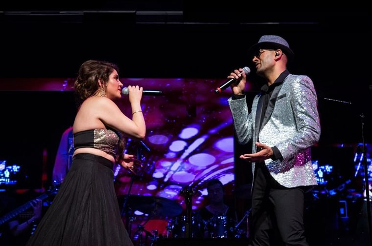 A man and woman on a stage facing each other. They are both holding microphones and singing. The man on the right is wearing a sequin silver blazer and hat, and the woman is wearing a long black skirt and a gold sequin top.
