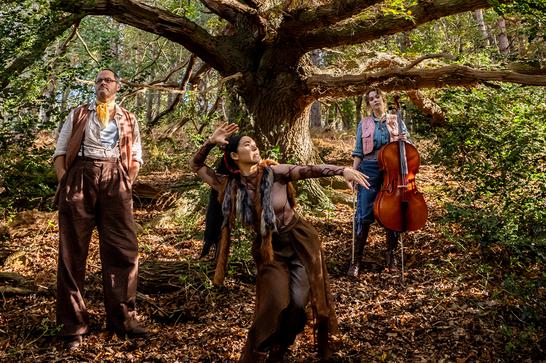 Three people stand in front of a large tree, one holding a cello to the right, one standing with their hands in their pocket and one expressively dancing in the centre