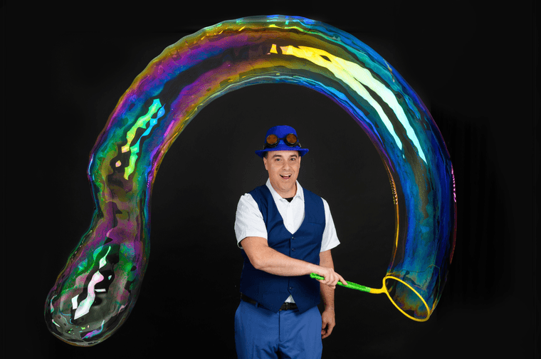 Man dressed in waist coat and bowler hat, blowing a large multicolour bubble that goes around his head.
