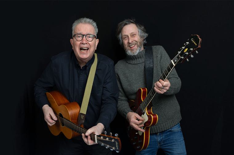 Two men smiling and playing the guitar. Man on left has short grey hair and wears glasses. Man on right has dark grey hair and a beard.