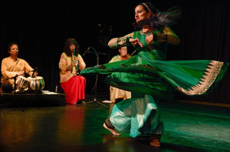 A woman in a green dress performs a traditional dance on stage while three musicians play instruments in the background.