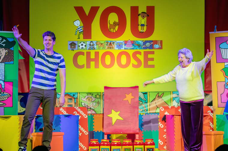 Man and woman stood on a stage. There are lots of colourful boxes and posters on the stage. In the middle of them is a large screen with the word 'You Choose' on it. The man is on the left and is wearing a blue and whte striped top. The woman is on the right and is wearing a yellow jumper and purple velvet trousers.