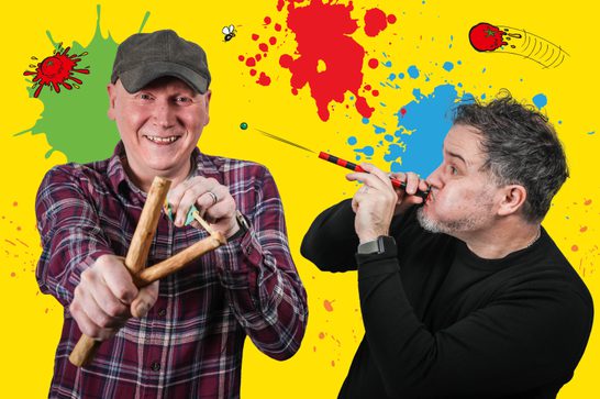 Two men against a colourful background. The man on the left wears a grey cap and red plaid shirt, smiling broadly while holding a wooden sling shot. The man on the right has curly grey hair and wears a black shirt, playing what appears to be a wooden flute or pipe with red and black striping.