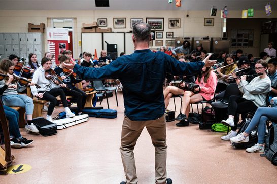 large group of children sat in a semi circle playing instruments like violin and clarinets. The children at the back of the group are playing guitars and drums. In the centre of the image is the back of a man who is facing the children. He has both arms out stretched and is conducting the musicians.