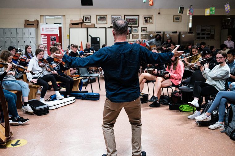 large group of children sat in a semi circle playing instruments like violin and clarinets. The children at the back of the group are playing guitars and drums. In the centre of the image is the back of a man who is facing the children. He has both arms out stretched and is conducting the musicians.