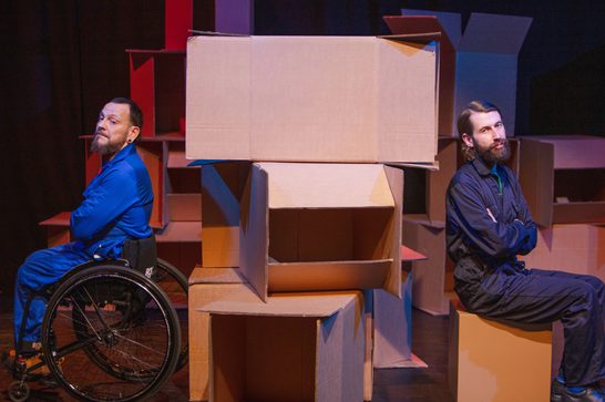 Jon, a white person with a beard wearing dark blue overalls and Daryl, a white man with a beard in light blue overalls, sit on either side of a wall of boxes with their backs to each other