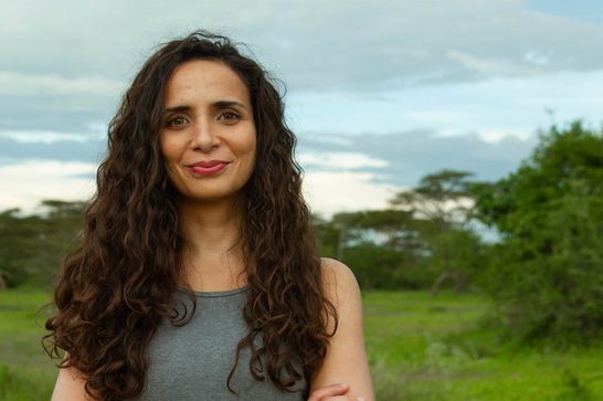 A woman with long, wavy brown hair wearing a grey sleeveless top smiles at the camera in an outdoor setting. Behind her is a lush green landscape with trees and grass under a cloudy sky.