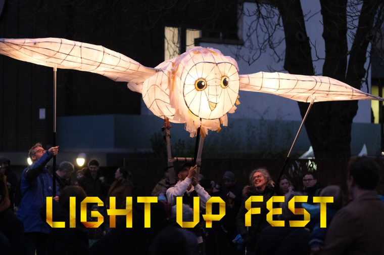 A large owl lantern with outstretched wings is carried through a crowd at Light Up Fest. The glowing paper lantern sculpture is held up by people using poles as spectators gather around.