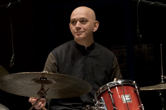 A drummer in a black button-up shirt sits behind a red drum kit, holding drumsticks over the cymbals. The photo is taken against a black background.