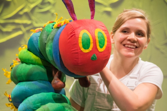 Woman holding a colourful handmade caterpillar puppet with a red head, green segmented body, and yellow accents.