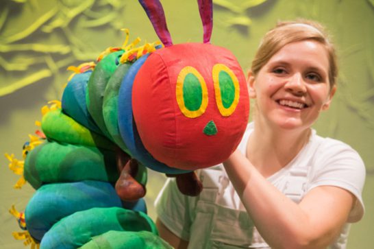 Woman holding a colourful handmade caterpillar puppet with a red head, green segmented body, and yellow accents.
