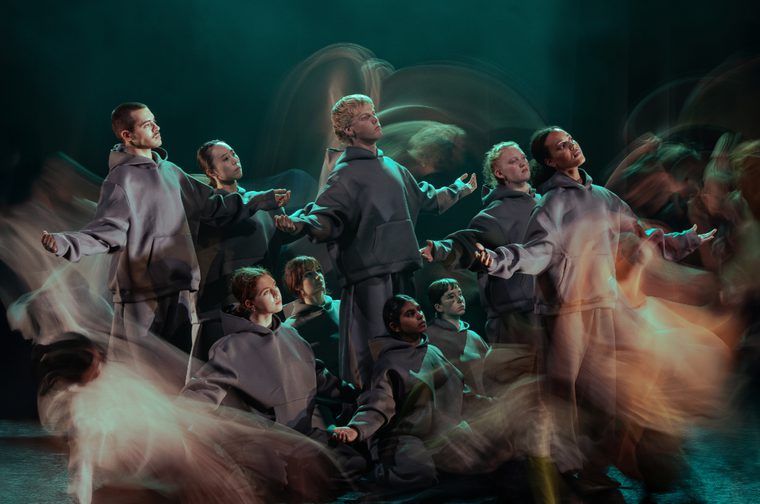 A dynamic long-exposure photograph of a dance or performance group wearing matching grey hooded sweatshirts against a teal-lit background. Multiple exposures create ghostly motion trails showing dancers in various poses with outstretched arms.